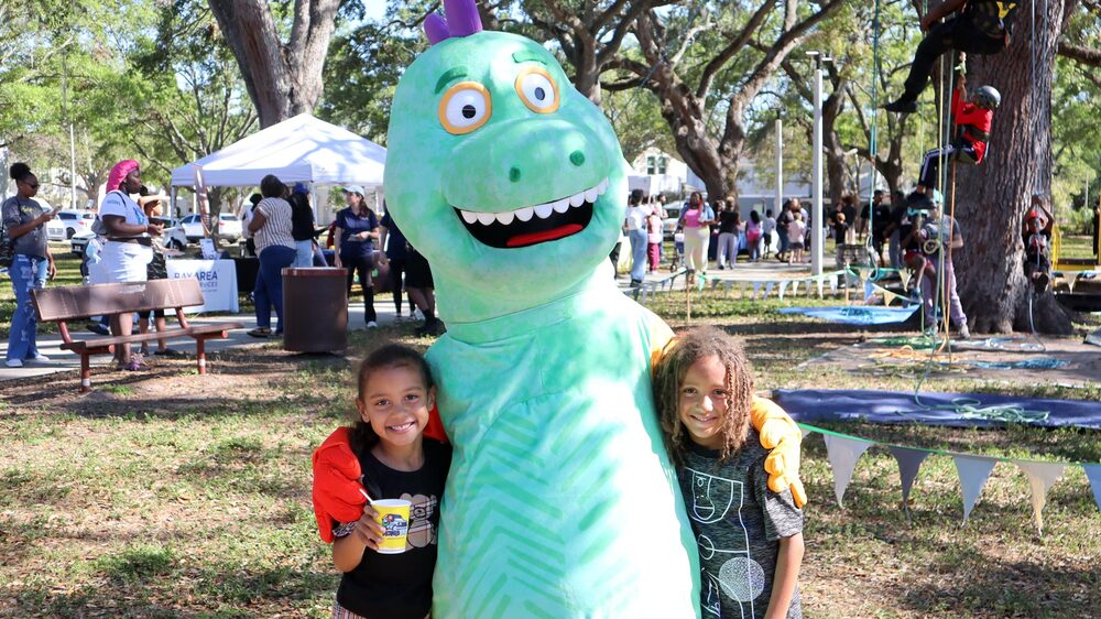 Two children with Great Explorations dinosaur mascot at Spring Family Picnic event in Jordan Park on March 18, 2025.