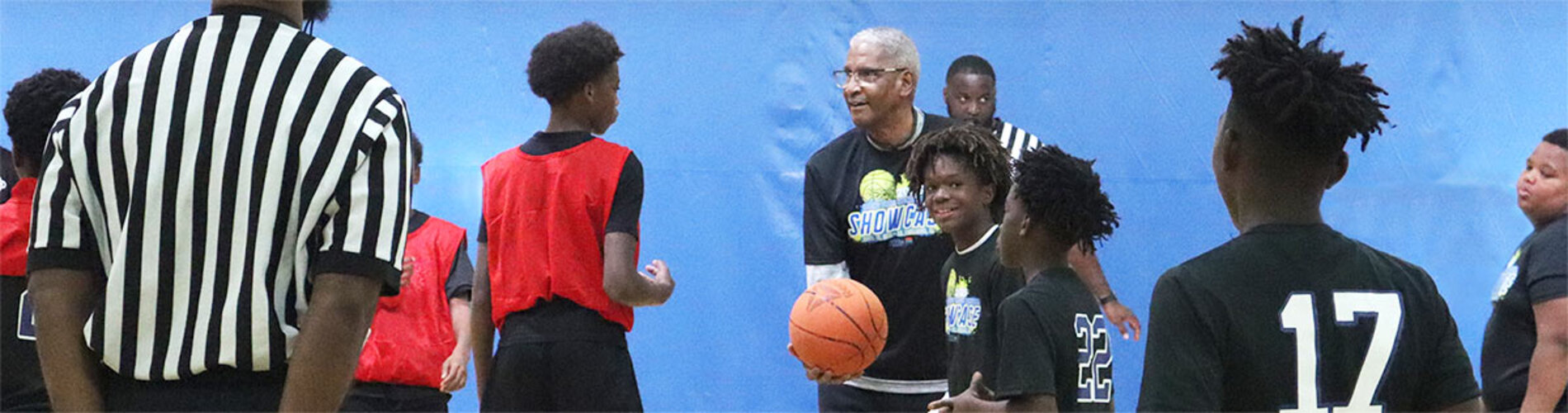 A basketball coach standing on a court holding a basket ball while taking to various children standing around the court.