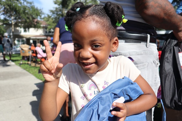 Young girl holding a backpack with her left hand and making the peace symbol hand gesture with her right hand.